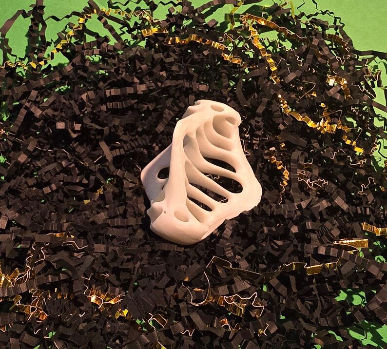Puede incluir: Objeto blanco de pl&aacute;stico impreso en 3D con un dise&ntilde;o esquel&eacute;tico, que se asemeja a una caja tor&aacute;cica, sobre un fondo de papel triturado negro y dorado.