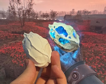 Op de afbeelding: Twee witte, getextureerde, eivormige objecten. Een object gloeit blauw en het andere is effen wit. De objecten worden in de handen van een persoon gehouden. De achtergrond is een veld met rode bloemen.