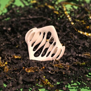Puede incluir: Objeto decorativo de pl&aacute;stico blanco con un dise&ntilde;o geom&eacute;trico calado. El objeto est&aacute; sentado en una cama de papel triturado negro y dorado.