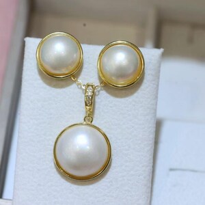 May include: A jewelry set consisting of two round stud earrings and a matching pendant, each featuring a large, smooth, white pearl set in a polished gold-tone metal frame. The pendant bail is accented with small, clear, sparkling stones.