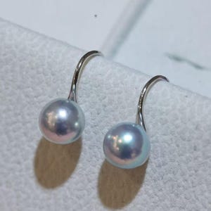 Könnte beinhalten: Ein Paar Ohrhänger mit glänzenden, kugelförmigen hellblauen Perlen, die an geschwungenen silberfarbenen Metallhaken hängen. Die Perlen haben eine glatte, reflektierende Oberfläche mit subtilen irisierenden Untertönen und sind an den Metallteilen befestigt.