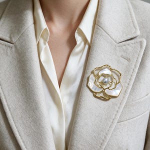 Könnte beinhalten: Eine goldfarbene Metallbrosche in Blumenform mit weißen Perlmuttblütenblättern und einer einzelnen zentralen Perle, befestigt an einem beigen Wollblazer über einem cremefarbenen Hemd mit Kragen.