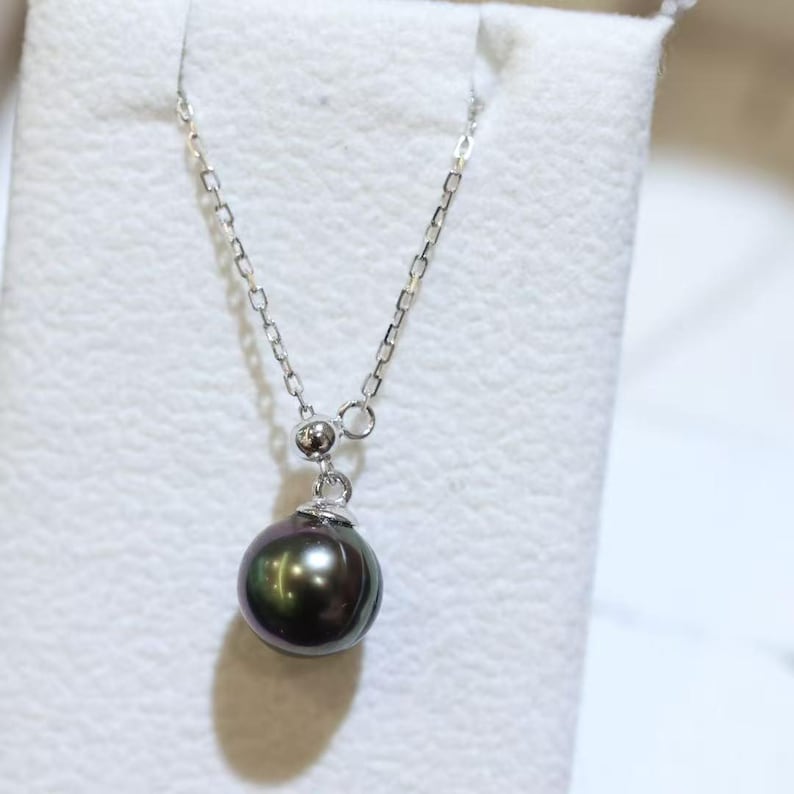 May include: A necklace featuring a single round, dark iridescent pearl pendant suspended from a delicate silver-toned metal chain. The pearl displays deep hues of black, green, and purple, secured by a small silver-toned metal cap and bail.