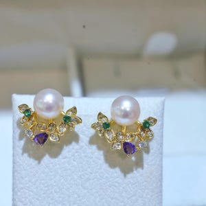Könnte beinhalten: Ein Paar goldene Ohrstecker mit einer zentralen weißen Perle, akzentuiert durch kleine grüne runde Steine, klare Kristalle und einen einzelnen violetten birnenförmigen Stein, die in einem floralen Fächermuster um die Perle angeordnet sind.