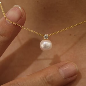 May include: A delicate gold necklace featuring a single, lustrous white pearl pendant. The pearl is accented by a small, sparkling clear gemstone. The necklace is held by a person.