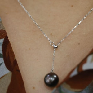 May include: A silver-toned Y-shaped necklace featuring a delicate chain and a single, smooth, dark gray spherical pearl pendant. The chain connects to a small circular ring, from which a shorter chain segment suspends the round, lustrous pearl.