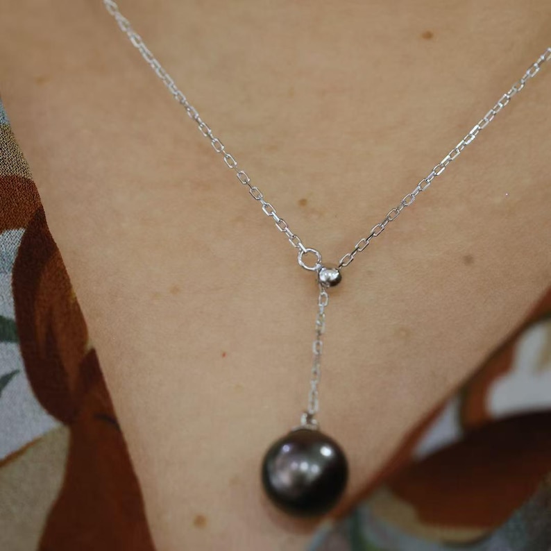 May include: A silver-toned Y-shaped necklace featuring a delicate chain and a single, smooth, dark gray spherical pearl pendant. The chain connects to a small circular ring, from which a shorter chain segment suspends the round, lustrous pearl.