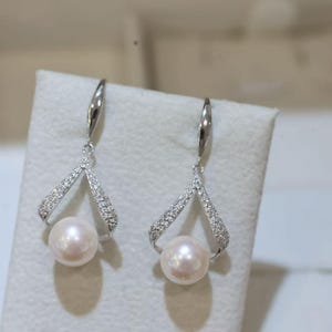 Könnte beinhalten: Ein Paar elegante Ohrhänger mit einer glänzenden, runden Perle, die in einem silberfarbenen, mit Diamanten besetzten Rahmen hängt. Die Ohrringe haben ein zartes, offenes Design und einen einfachen Hakenverschluss. Die Perlen haben eine weiche, cremefarbene Farbe.
