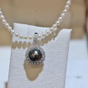 May include: A pearl necklace with a dark gray pearl pendant. The pendant is surrounded by a halo of small, clear gemstones. The necklace features a strand of small, white pearls.