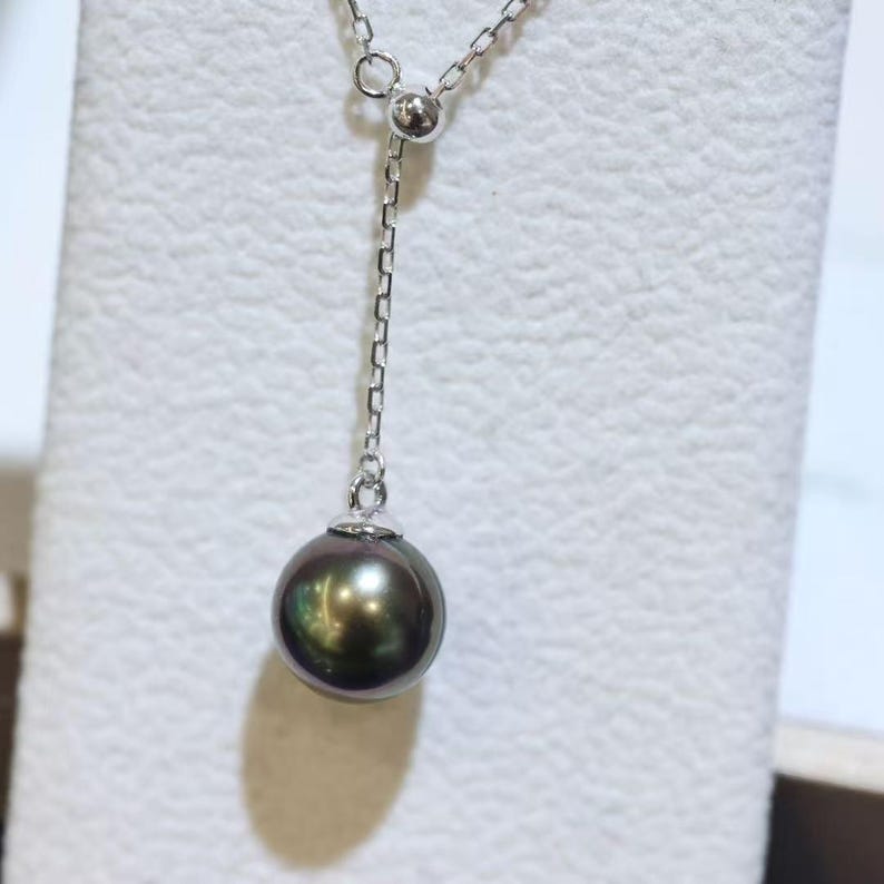 May include: A lariat-style necklace featuring a single round, dark iridescent pearl pendant suspended from a delicate silver-toned chain. The chain includes a small, polished silver-toned bead accent where the drop section connects to the main chain.