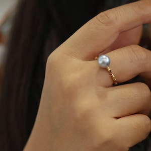 Könnte beinhalten: Ein goldener Ring mit einer großen, runden, grauen Perle. Der Ring hat ein zartes, strukturiertes Band. Die Perle ist der Blickfang, prominent auf dem Finger platziert.