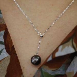 May include: A silver-toned lariat necklace featuring a delicate cable chain and a single dark, lustrous spherical pearl pendant suspended from a central circular link. The chain extends downward from the center point to hold the round dark pearl.