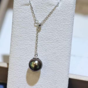 May include: A silver lariat necklace featuring a delicate chain and a single, round, dark iridescent pearl pendant. The chain passes through a small silver bead, allowing the length of the drop to be adjusted, creating a sleek and polished look.