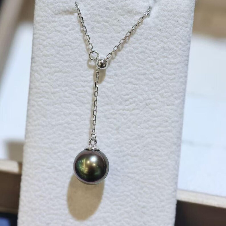 May include: A silver lariat necklace featuring a delicate chain and a single, round, dark iridescent pearl pendant. The chain passes through a small silver bead, allowing the length of the drop to be adjusted, creating a sleek and polished look.