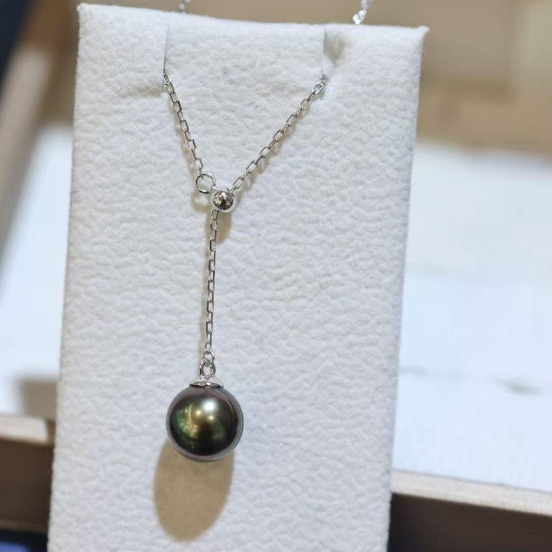 May include: A silver-toned chain necklace featuring a single dark, iridescent pearl pendant. The chain passes through a small, round silver-toned bead, allowing the pearl to hang at the end of a vertical drop section of the delicate metal chain.
