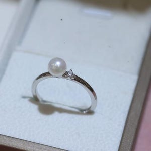 Könnte beinhalten: Ein silberner Ring mit einer runden Perle und einem kleinen, funkelnden Akzentstein. Der Ring hat ein zartes, minimalistisches Design, das sich für den täglichen Gebrauch eignet. Der Ring wird in einer Schmuckschatulle präsentiert.