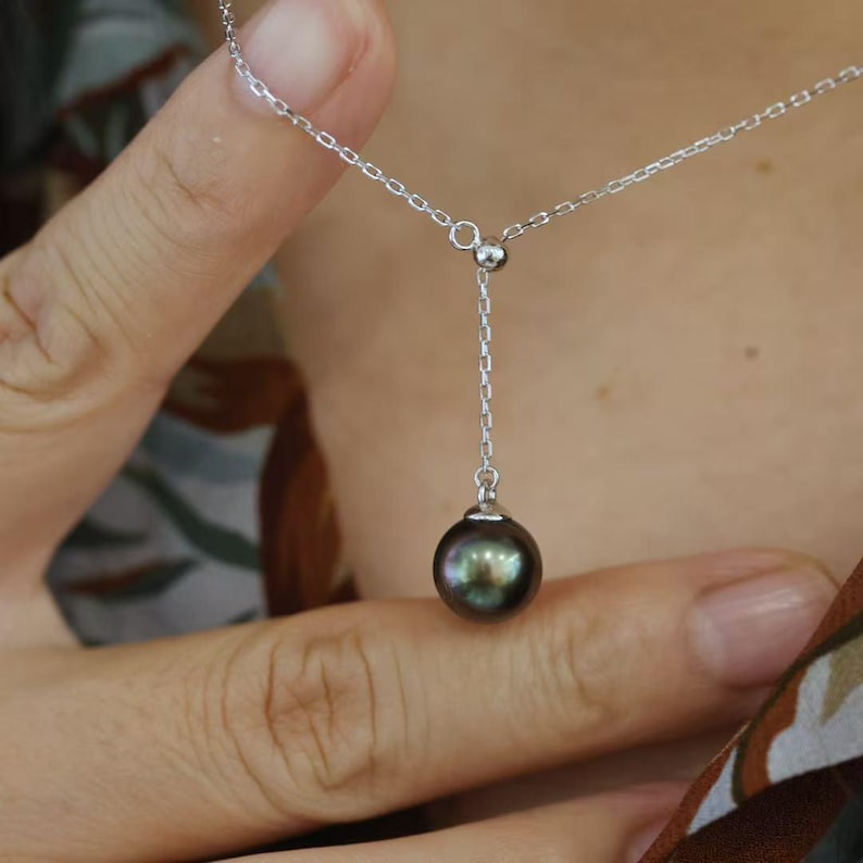 May include: A silver chain necklace featuring a single dark, iridescent pearl pendant. The chain forms a Y-shape, with a delicate silver link design connecting to a small metal bead that holds the round, dark-colored pearl at the bottom center.