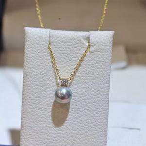 May include: A gold-colored chain necklace with a pendant featuring a round, iridescent pearl. The pearl is suspended below a small, square, diamond-studded setting. The necklace is displayed on a white, textured surface.