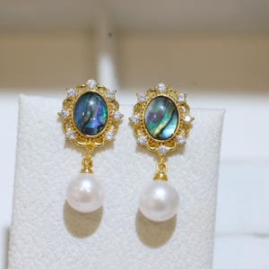 Könnte beinhalten: Ein Paar goldfarbene Ohrringe mit einem ovalen, irisierenden Abalone-Muschel-Cabochon, umgeben von einem dekorativen Filigranrahmen mit kleinen klaren Kristallen und einer weißen, kugelförmigen Perle, die an der Basis hängt.