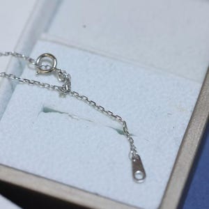 May include: A silver-toned metal chain necklace featuring small, uniform rectangular links. The chain includes a circular spring ring clasp mechanism and a flat, elongated metal tag at the end, showcasing a polished, reflective metallic finish.
