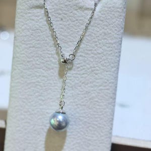 May include: A delicate silver necklace featuring a single, iridescent gray pearl pendant. The chain is fine and elegant, with a simple design that highlights the pearl's natural beauty. The necklace is displayed on a white background.
