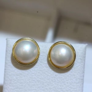 Könnte beinhalten: Ein Paar Ohrstecker mit Perlen. Jeder Ohrring hat eine große, glänzende, cremefarbene Perle, die in eine goldfarbene Fassung gefasst ist. Die Perlen haben einen weichen, schillernden Glanz, und die goldene Fassung sorgt für einen klassischen, eleganten Look.