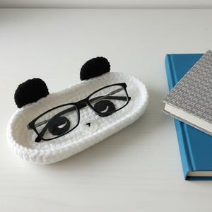Könnte beinhalten: Ein weißes, gehäkeltes Tablett in Pandaform mit schwarzen Ohren und einem Pandagesicht, das eine Brille mit schwarzem Rahmen hält. Ein blaues Buch und ein grau gemustertes Buch liegen neben dem Tablett auf einer weißen Oberfläche.