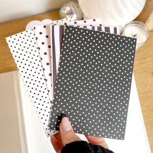 May include: A hand holds a stack of patterned cards in black and white. The cards feature polka dots and stripes. A notebook with a gold accent is visible in the background.