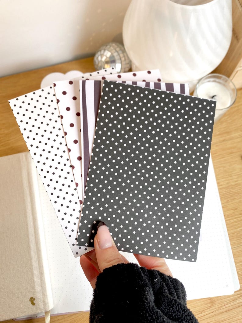 May include: A hand holds a stack of patterned cards in black and white. The cards feature polka dots and stripes. A notebook with a gold accent is visible in the background.