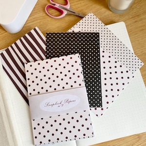 May include: A collection of scrapbook papers with various patterns, including polka dots and stripes, in shades of brown and black. The papers are bundled together with a label that reads "Scrapbook Papers n&deg;3". A pair of pink-handled scissors and a candle are also visible.