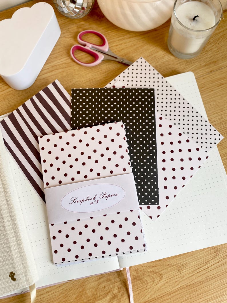 May include: A collection of scrapbook papers with various patterns, including polka dots and stripes, in shades of brown and black. The papers are bundled together with a label that reads "Scrapbook Papers n&deg;3". A pair of pink-handled scissors and a candle are also visible.