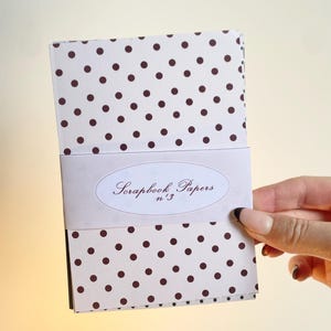 May include: A stack of scrapbook papers with a white background and brown polka dots. A white band with the text "Scrapbook Papers n&deg;3" wraps around the papers. The papers are held by a hand.