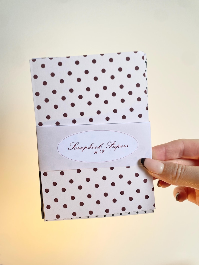 May include: A stack of scrapbook papers with a white background and brown polka dots. A white band with the text "Scrapbook Papers n&deg;3" wraps around the papers. The papers are held by a hand.