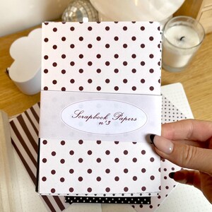 May include: A stack of scrapbook papers with a white background and brown polka dots. The papers are wrapped with a white band that reads "Scrapbook Papers n&deg;3" in elegant script. Additional patterned papers are visible.