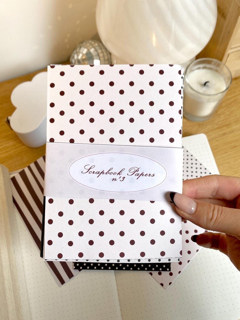 May include: A stack of scrapbook papers with a white background and brown polka dots. The papers are wrapped with a white band that reads "Scrapbook Papers n&deg;3" in elegant script. Additional patterned papers are visible.