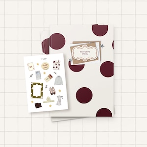 May include: A cream-colored notebook with large dark red polka dots, featuring a brown envelope with a label reading Souvenirs Diary. Beside it is a white sticker sheet displaying various illustrations including a frame, shoes, and a coffee pot.
