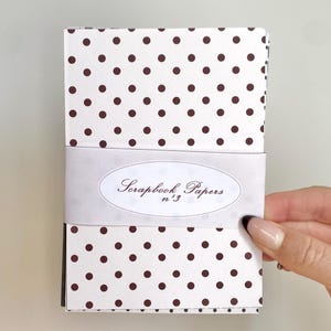 May include: A stack of scrapbook papers wrapped with a white band. The papers have a white background with a repeating pattern of dark brown polka dots. The band features the text "Scrapbook Papers n.3" in an oval design.