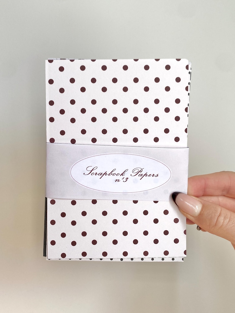 May include: A stack of scrapbook papers wrapped with a white band. The papers have a white background with a repeating pattern of dark brown polka dots. The band features the text "Scrapbook Papers n.3" in an oval design.