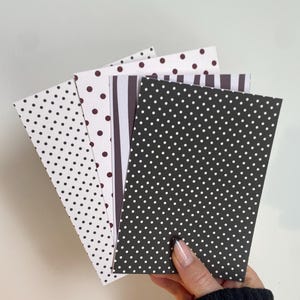 May include: A hand holding a stack of patterned cards. The cards feature polka dots, stripes, and solid colors in black, white, and brown. The cards are rectangular and appear to be made of paper.