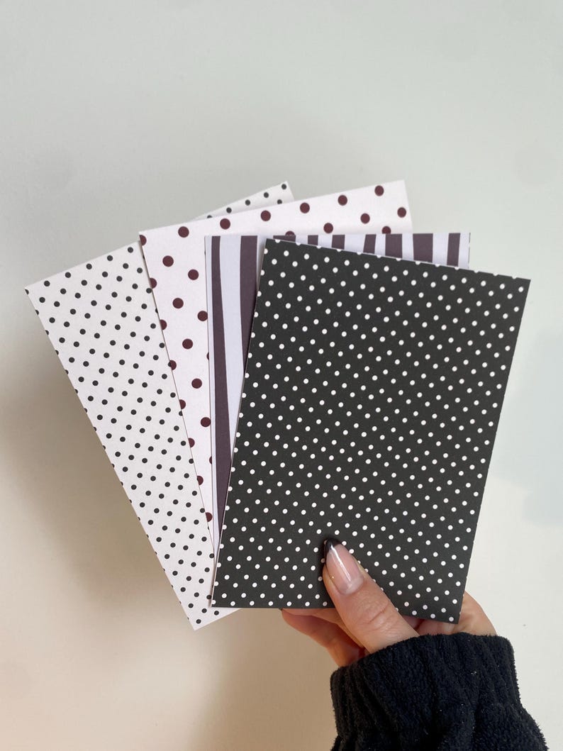 May include: A hand holding a stack of patterned cards. The cards feature polka dots, stripes, and solid colors in black, white, and brown. The cards are rectangular and appear to be made of paper.