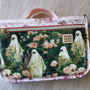 May include: A rectangular quilted zipper pouch with a top handle, featuring a dark green fabric printed with white ghosts and pink roses. A rectangular label on the front reads ROCK BABY SCISSORS. The pouch has a pink floral border and a metal zipper.