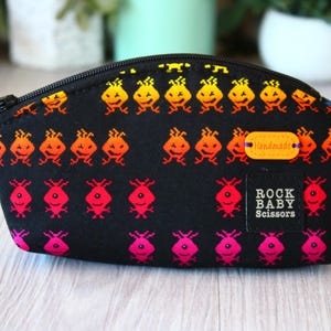 Könnte beinhalten: Schwarze Reißverschlusstasche mit einem pixeligen Alien-Muster in Gelb, Orange und Pink. Die Tasche hat ein kleines "handmade"-Etikett und ein "Rock Baby Scissors"-Etikett.
