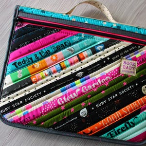 May include: A colorful, quilted fabric bag with a zipper closure and a small handle. The bag features diagonal stripes of various colors and patterns, including text like "Teddy & the Bears" and "Ruby Star Society". A small label reads "Rock Baby Soissons".