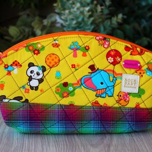 May include: A yellow quilted cosmetic bag with a colorful cartoon print featuring a panda, elephant, and other whimsical characters. The bag has an orange zipper and a plaid bottom. The label reads "ROCK BABY Scissors".