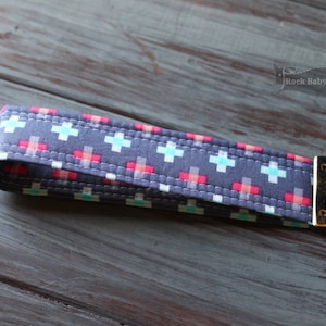 May include: A gray fabric key chain with a white and turquoise cross pattern and red accents. The key chain has a gold metal clasp.