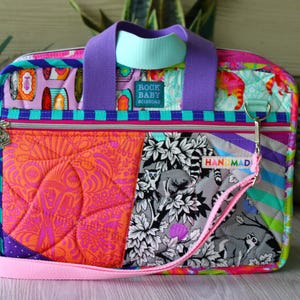 May include: A rectangular quilted laptop bag featuring a vibrant patchwork design with orange, purple, and black and white lemur patterns. It includes purple handles, a light blue strap, a front zipper pocket, and a label reading ROCK BABY SCISSORS.