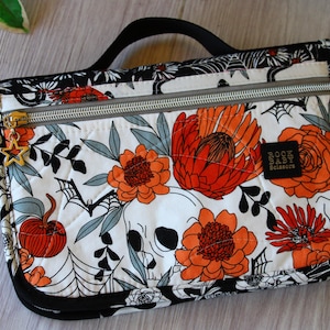 May include: A quilted, zippered pouch with a black handle. The white fabric is patterned with orange flowers, gray leaves, a skull, bats, and spiderwebs. A gold star charm hangs from the zipper. The label reads "Rock Baby Scissors."