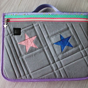 May include: Gray quilted laptop sleeve with a pink and blue star design. The sleeve has a purple trim, a zipper with a star charm, and a handle. The design includes a colorful stripe near the zipper.