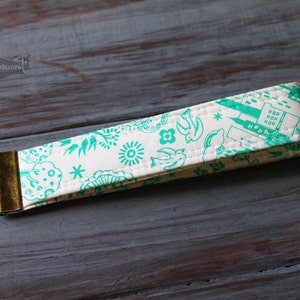 May include: A white key fob featuring a teal pattern of birds, flowers, and buildings. The key fob has a brass-coloured metal loop and is made of a quilted fabric. The key fob is on a grey wooden surface.