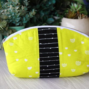 May include: A bright yellow cosmetic bag with a white zipper and a black vertical stripe. The bag is decorated with white floral patterns. The bag is made of fabric and is designed to hold makeup and other beauty products.